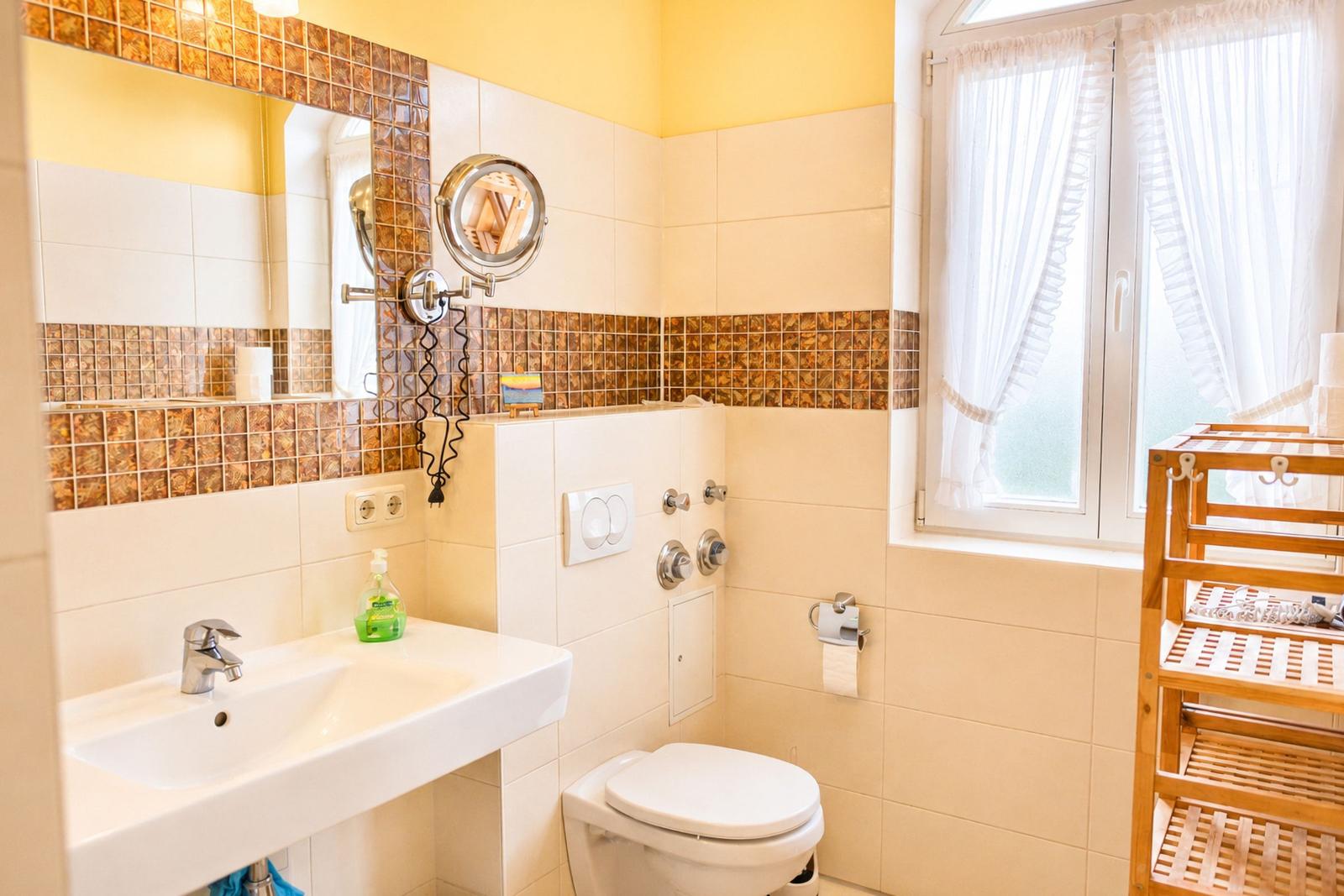 Badezimmer mit Waschbecken, Toilette und Dusche. Fenster mit weißen Vorhängen.