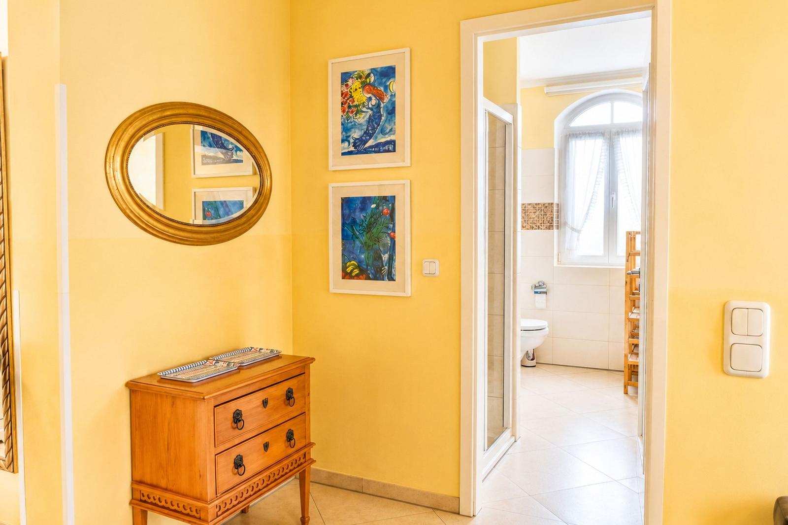 Helle Wand mit Spiegel, Schrank und zwei Bildern. Tür führt in Bad mit Toilette.