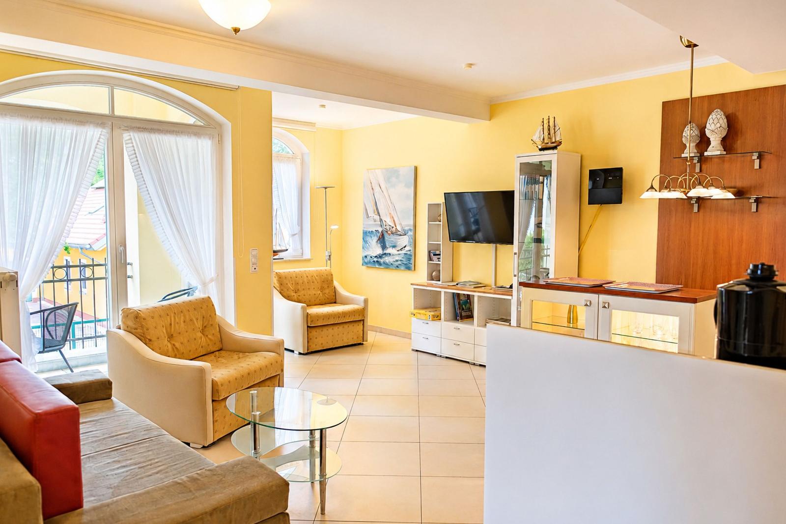 Wohnzimmer mit gelben Wänden, Sitzmöbeln und TV. Balkon mit Türen. Wandbild und Schrank.