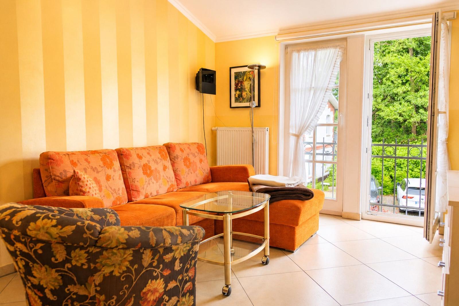 Wohnzimmer mit orangefarbenem Sofa, Glas-Tisch und Balkontür zu grünem Außenbereich.