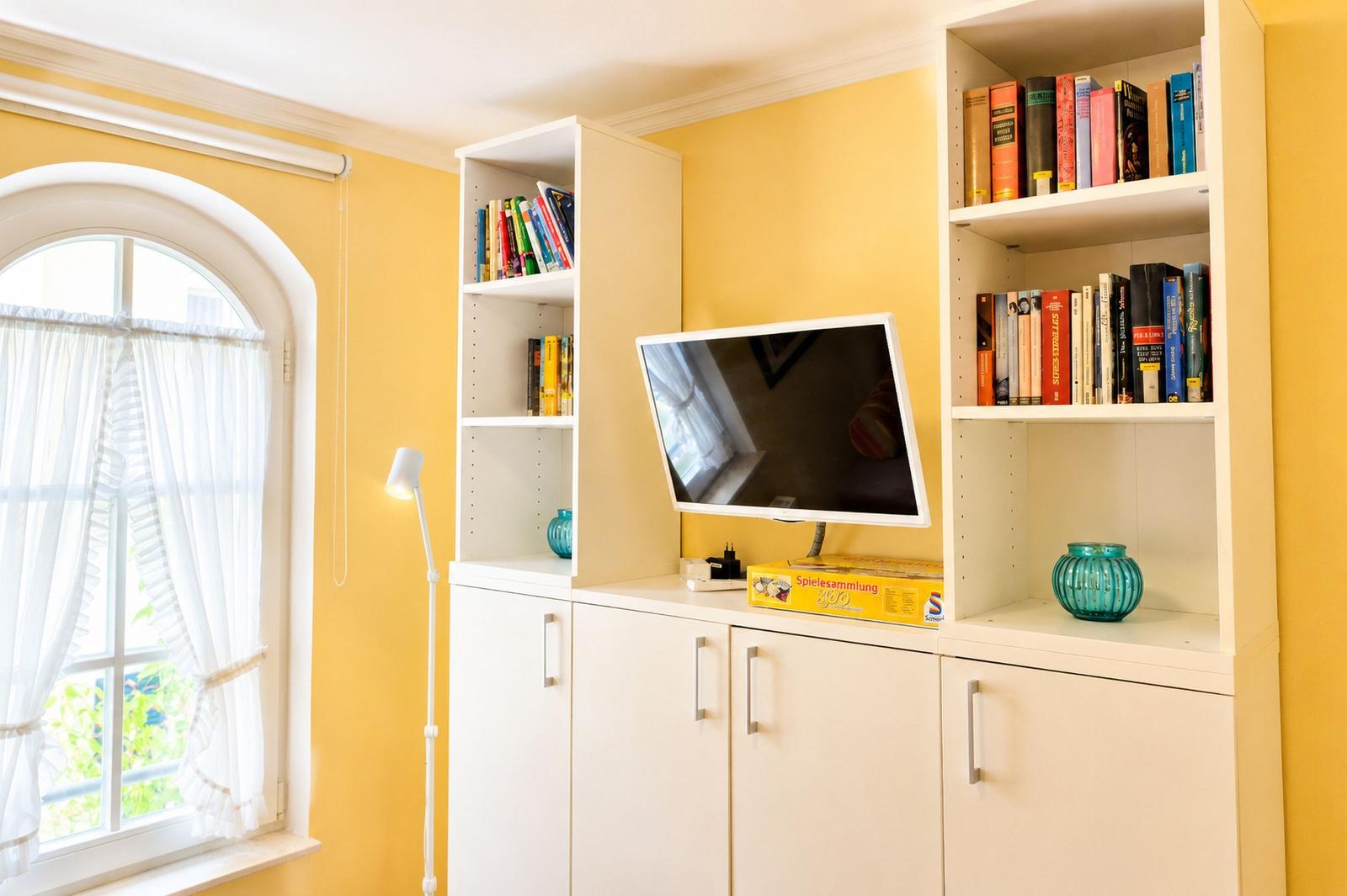 Ein Raum mit gelben Wänden, einem weißen Bücherregal, einem Fernseher und einem Fenster mit weißen Vorhängen.