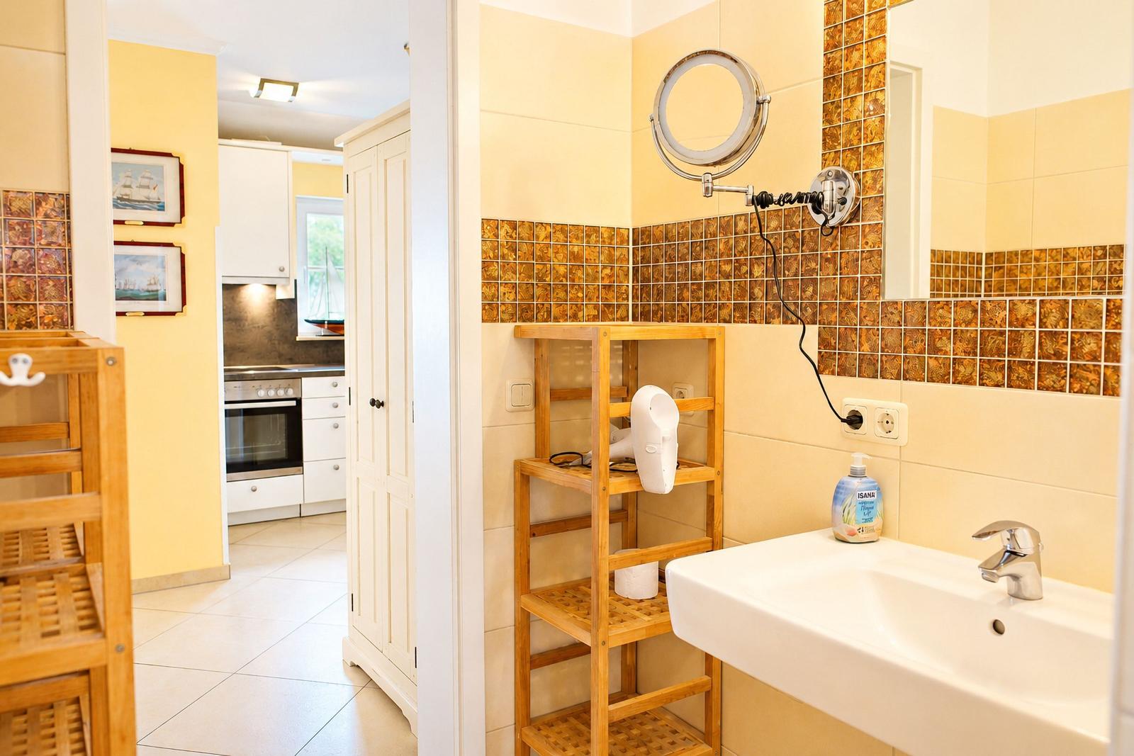 Badezimmer mit Waschbecken, Spiegel und Holzregal. Küche im Hintergrund sichtbar.