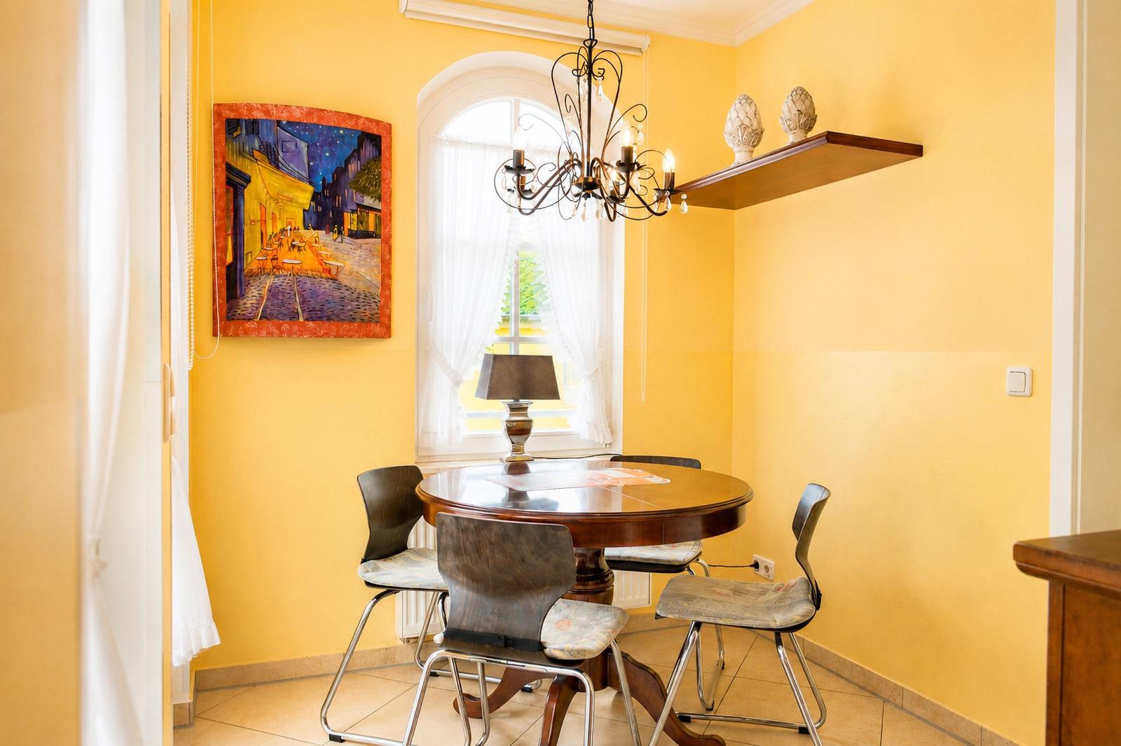 Esszimmer mit runden Tisch, vier Stühlen, Wandbild und Kronleuchter.
