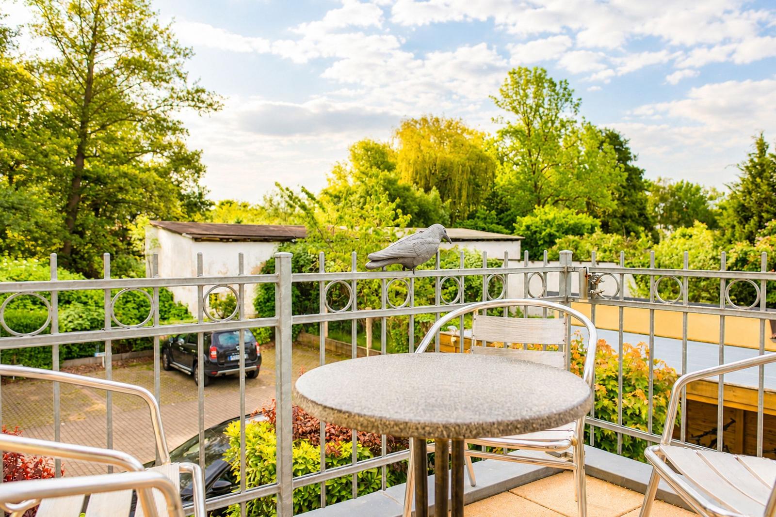 Terrasse mit Tisch und Stühlen, Vogelstatue auf Geländer, Blick auf Grünflächen und Hof.