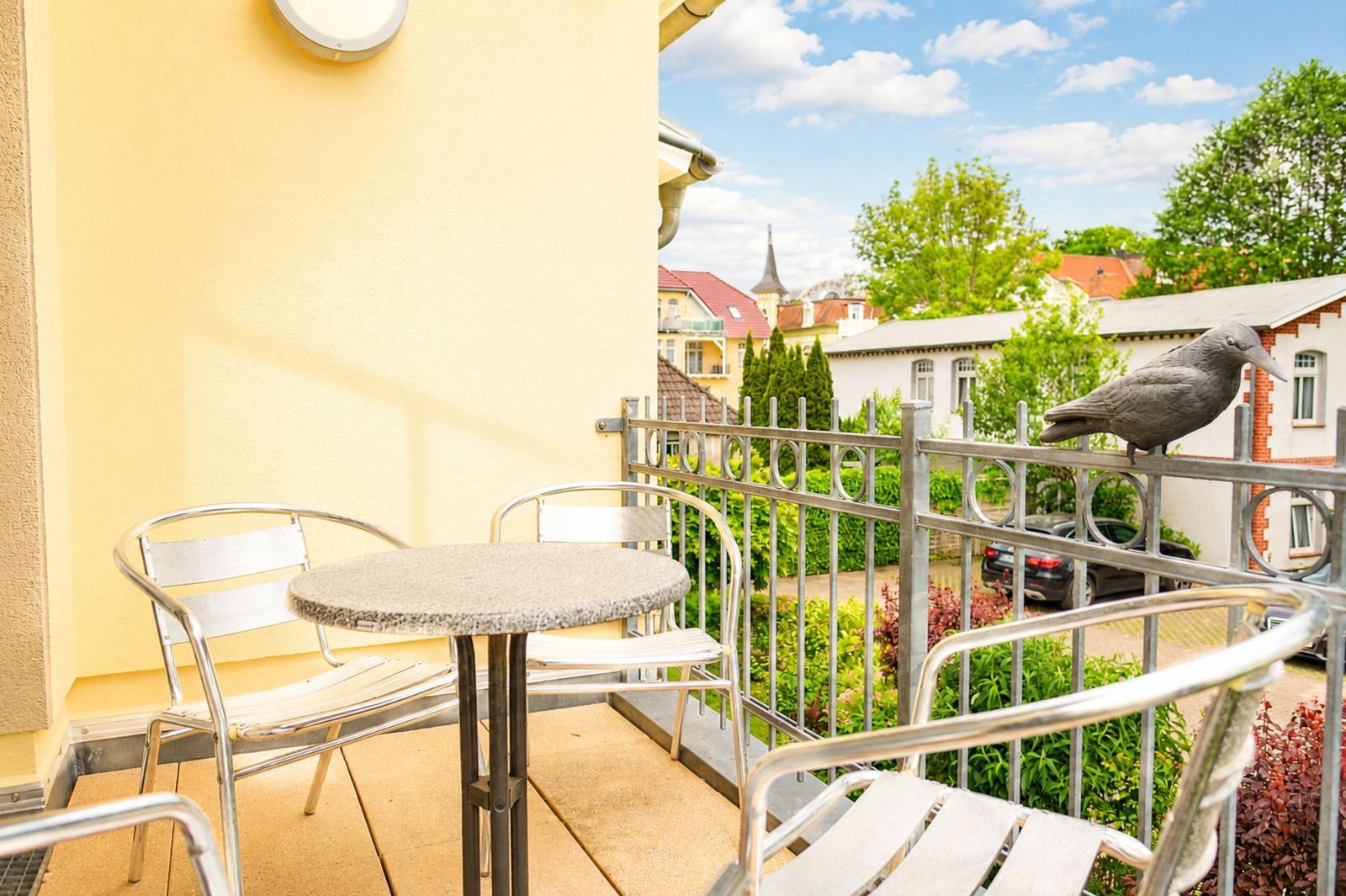 Terrasse mit Tisch und Stühlen, Vogelskulptur auf Geländer, Blick auf Wohngebiet mit Bäumen.