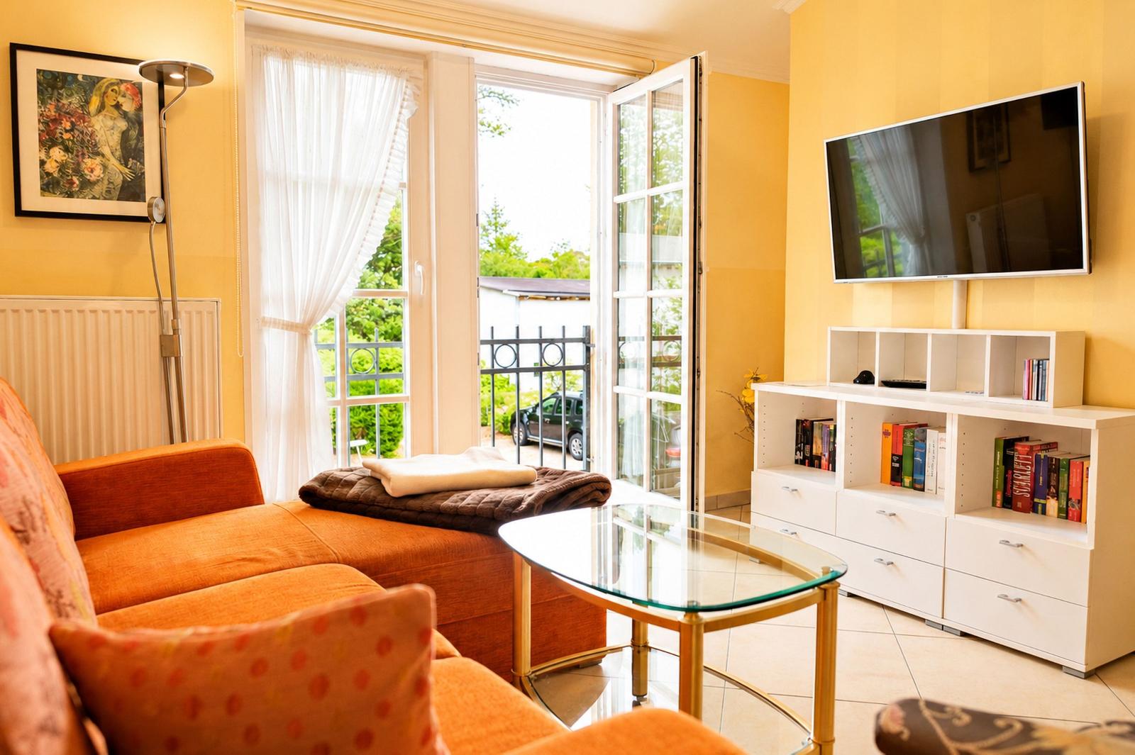 Wohnzimmer mit orangefarbenem Sofa, Glas-Tisch und Fernseher an der Wand.