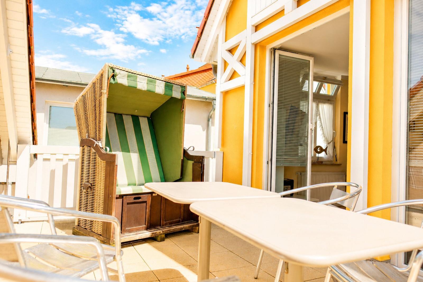 Terrasse mit Strandkorb, Tischen und Stühlen vor gelbem Haus mit Schiebetür.