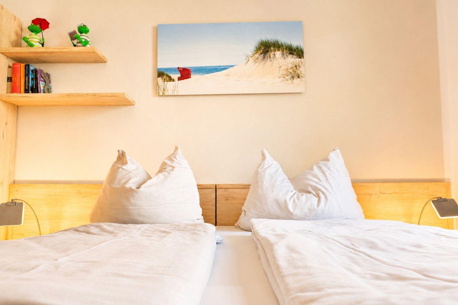 Zwei Betten mit weißen Laken und Kissen. Wandbild zeigt Strand. Holzregale mit Büchern und Deko.