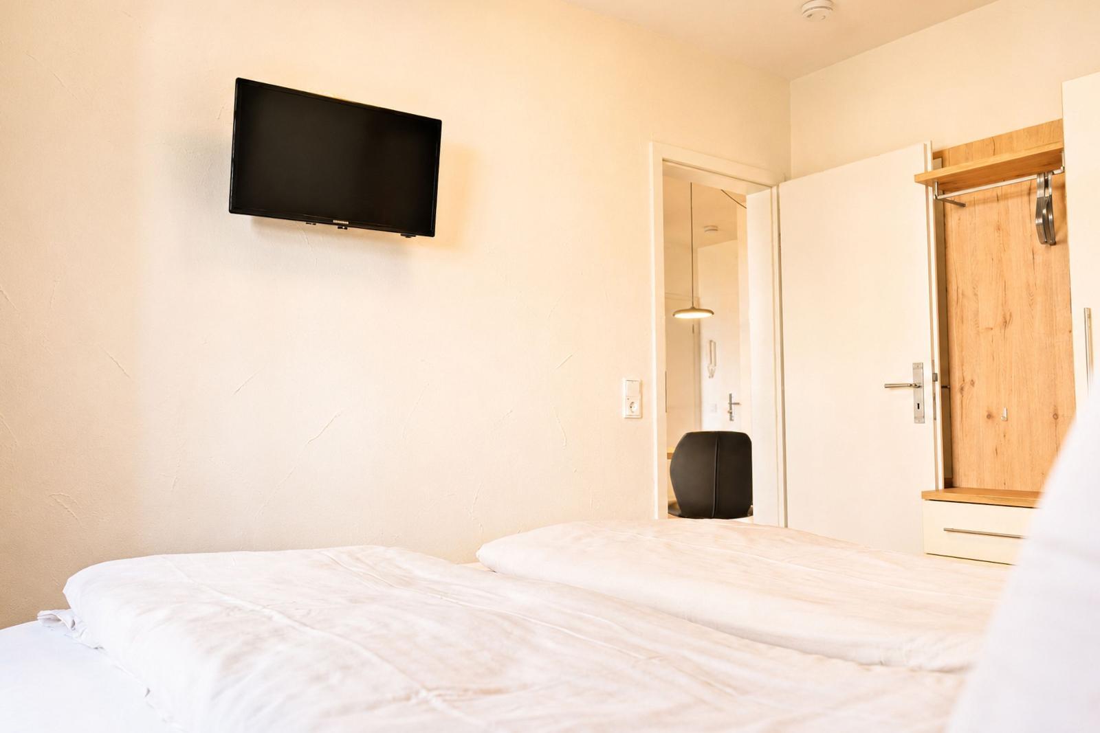 Schlafzimmer mit Bett, Wand-TV und Kleiderschrank. Tür führt in angrenzenden Raum.