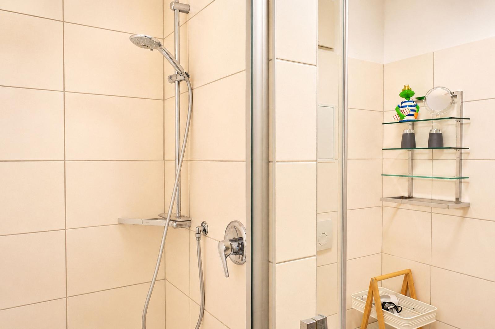 Dusche mit Glaswand, Duschbrause und Ablage mit Kosmetikartikeln.