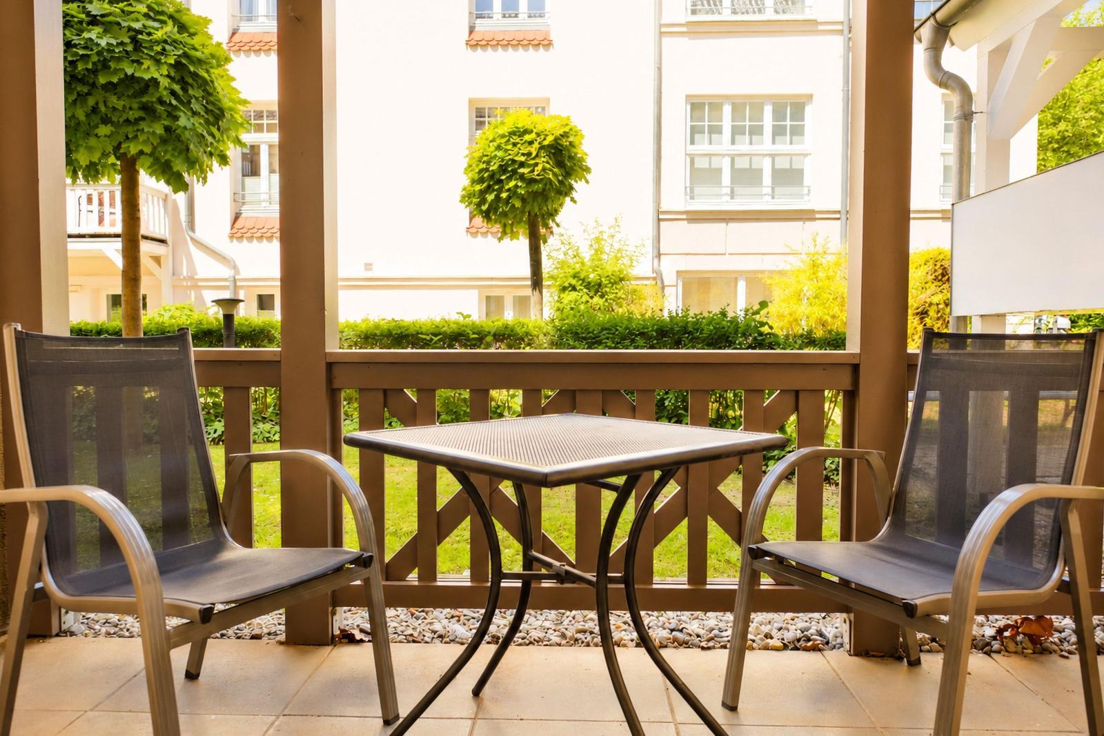 Terrasse mit Tisch und zwei Stühlen, Blick auf grünen Garten und Gebäude.