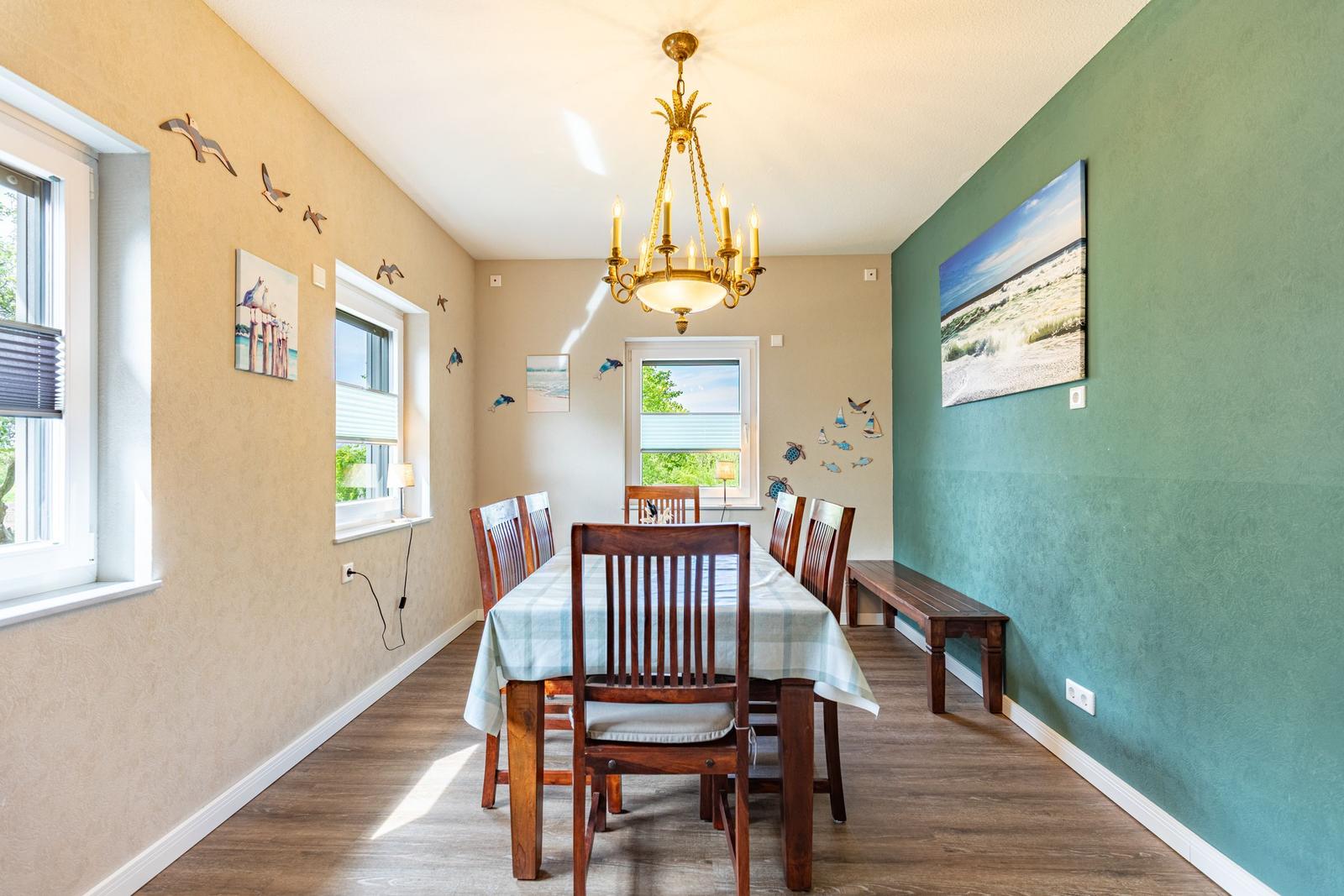 Esszimmer mit Holztisch, Stühlen und Wandbildern. Deckenleuchte und Fenster sichtbar.