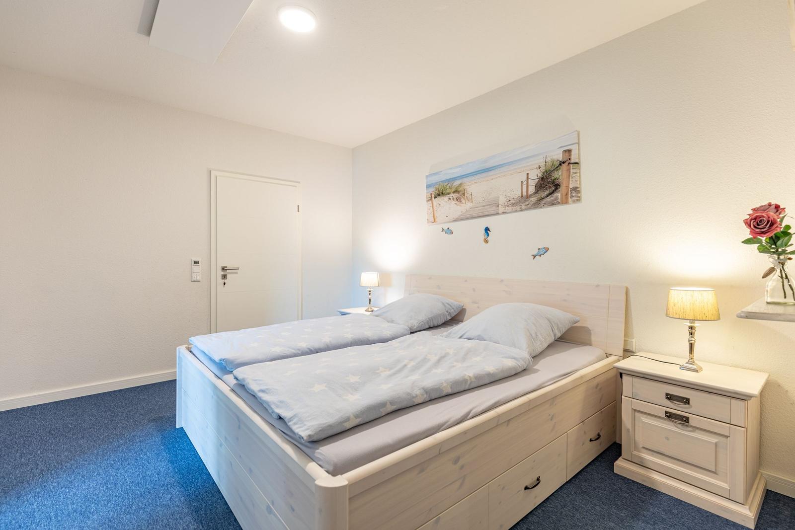 Schlafzimmer mit Doppelbett, Nachttisch und Wandbild. Blauer Teppich und weiße Wände.