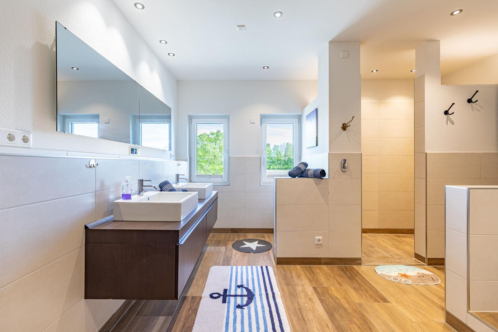 Badezimmer mit zwei Waschbecken, Dusche und Holzboden. Anker-Teppich und Fenster mit Blick auf Grün.