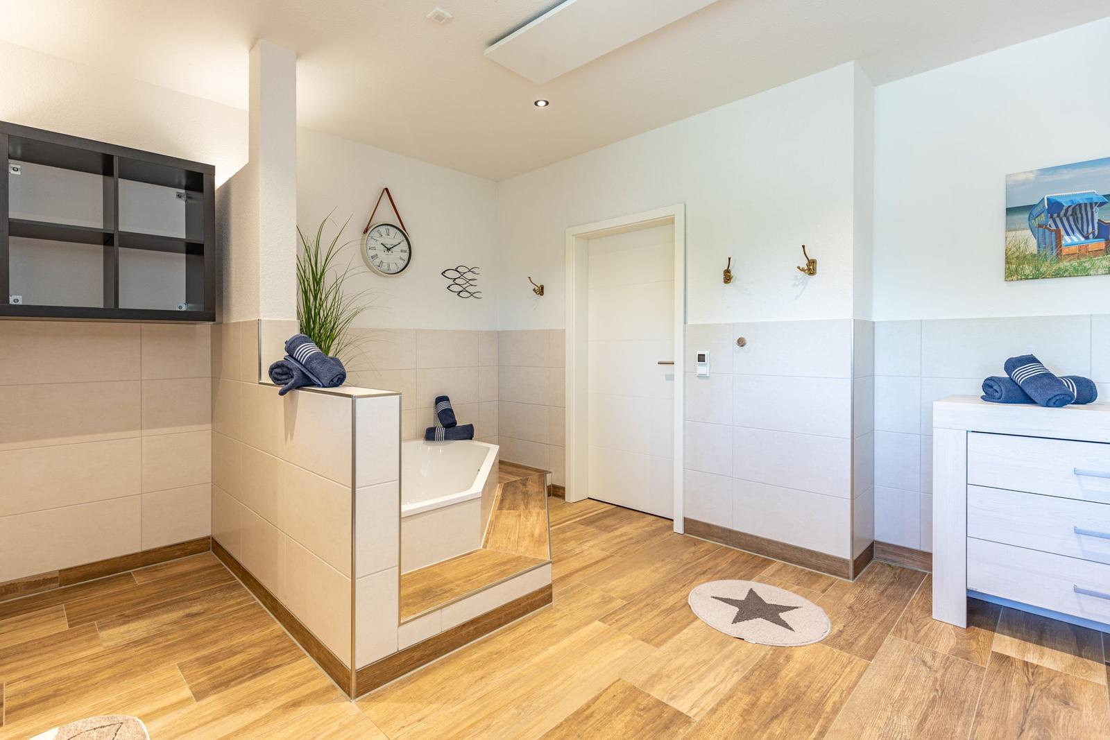 Bad mit Badewanne, Holzfußboden und Schrank. Wanduhr und Strandbild an der Wand.