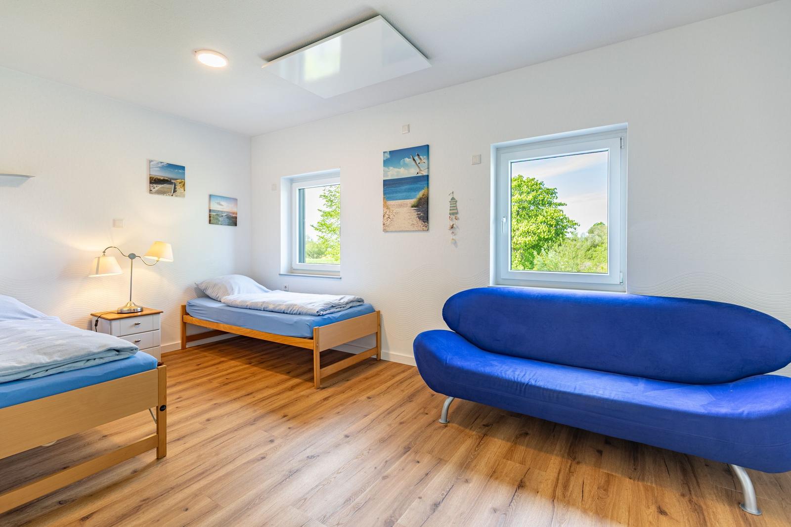 Zimmer mit zwei Betten, blauem Sofa und Holzboden. Fenster zeigen grüne Bäume.
