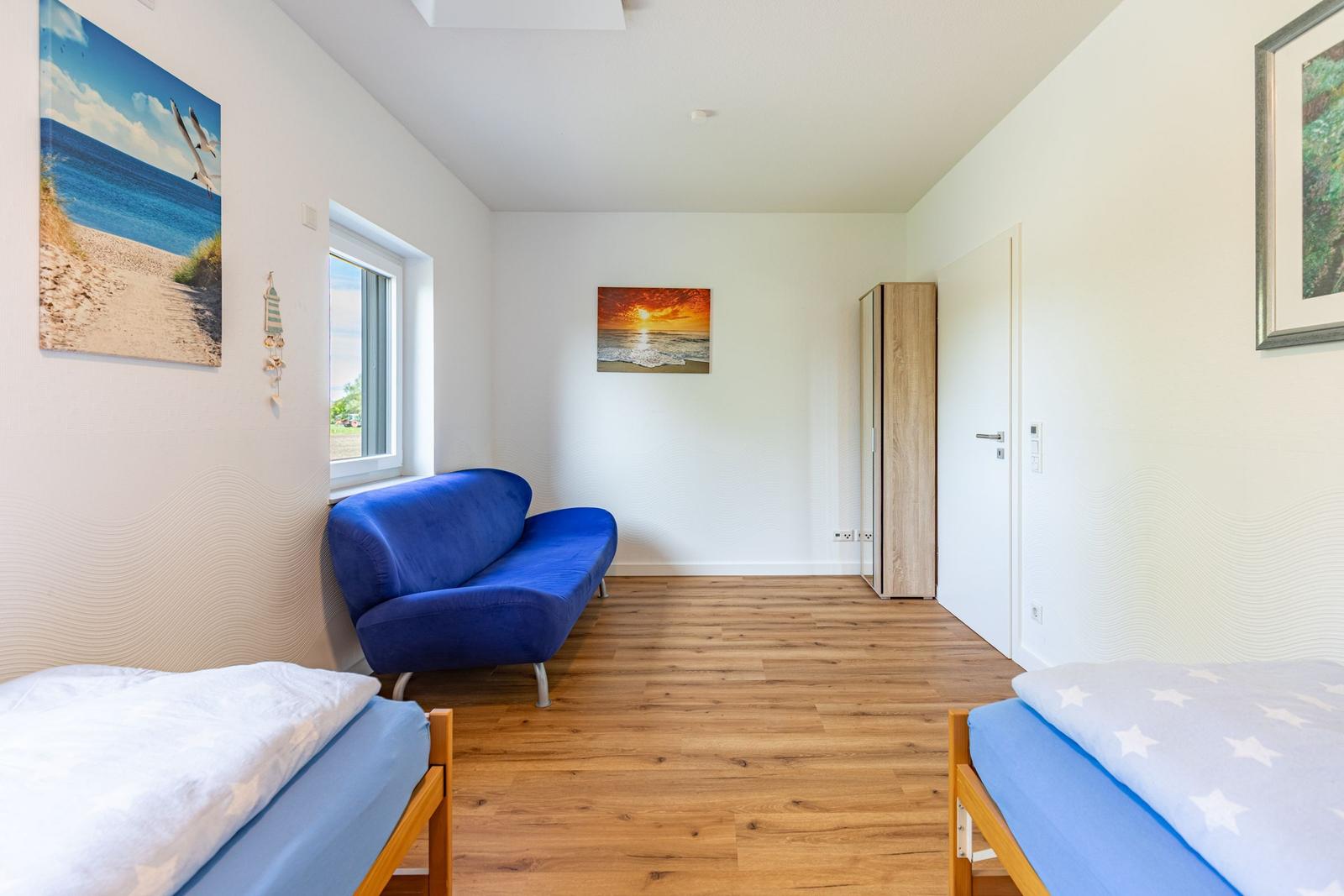 Zimmer mit zwei Betten, blauem Sofa und Holzboden. Wandbilder zeigen Meereslandschaften.