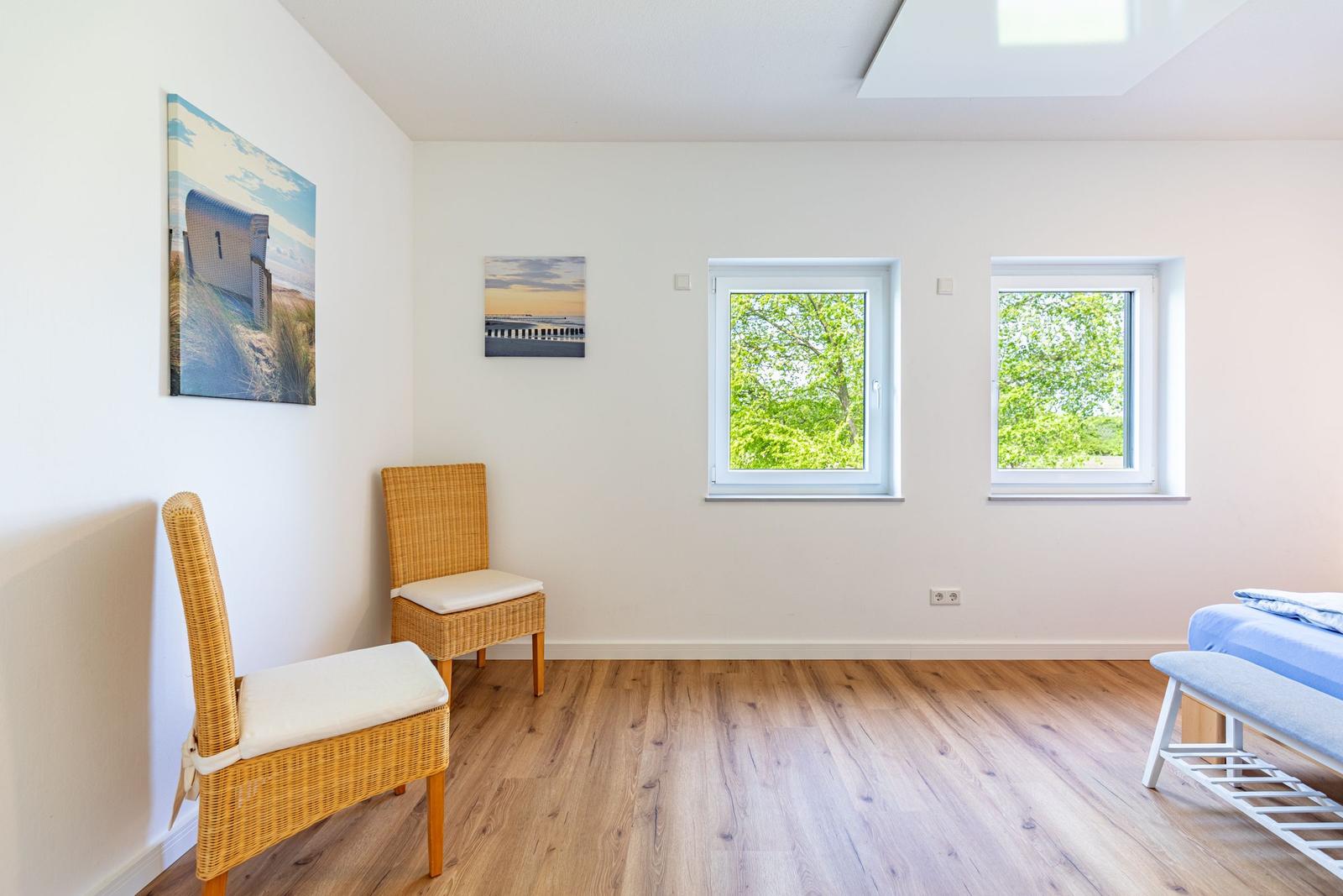 Zimmer mit zwei Fenstern, Holzboden und zwei Stühlen. Wandbilder und Bett sind sichtbar.