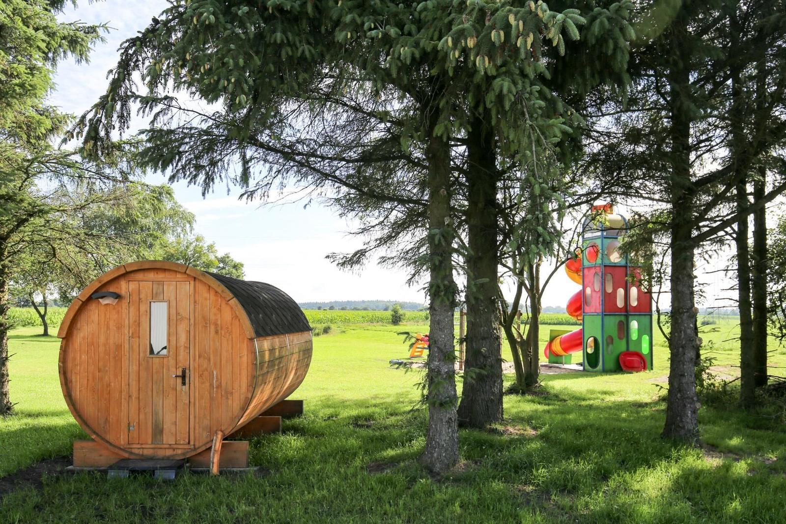 Holzsauna und Kinderbereich unter Bäumen im Gras.