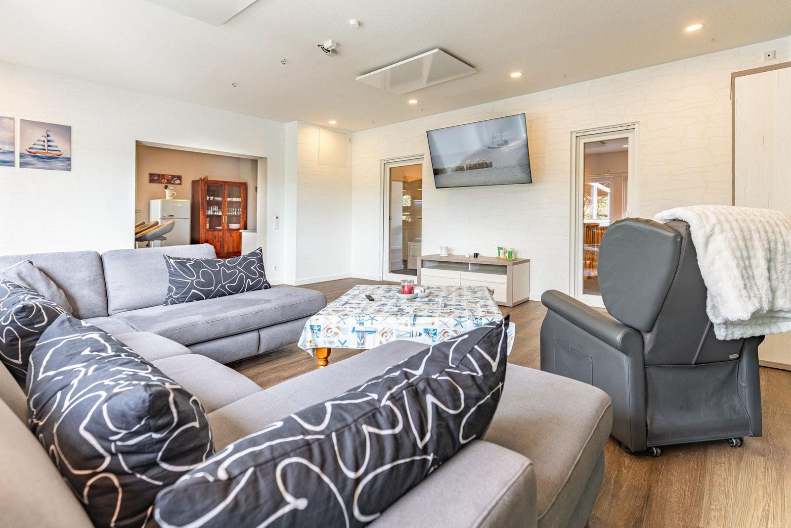 Wohnzimmer mit Sofa, Fernseher und Sessel. Tisch mit Tischdecke und Bilder an der Wand.