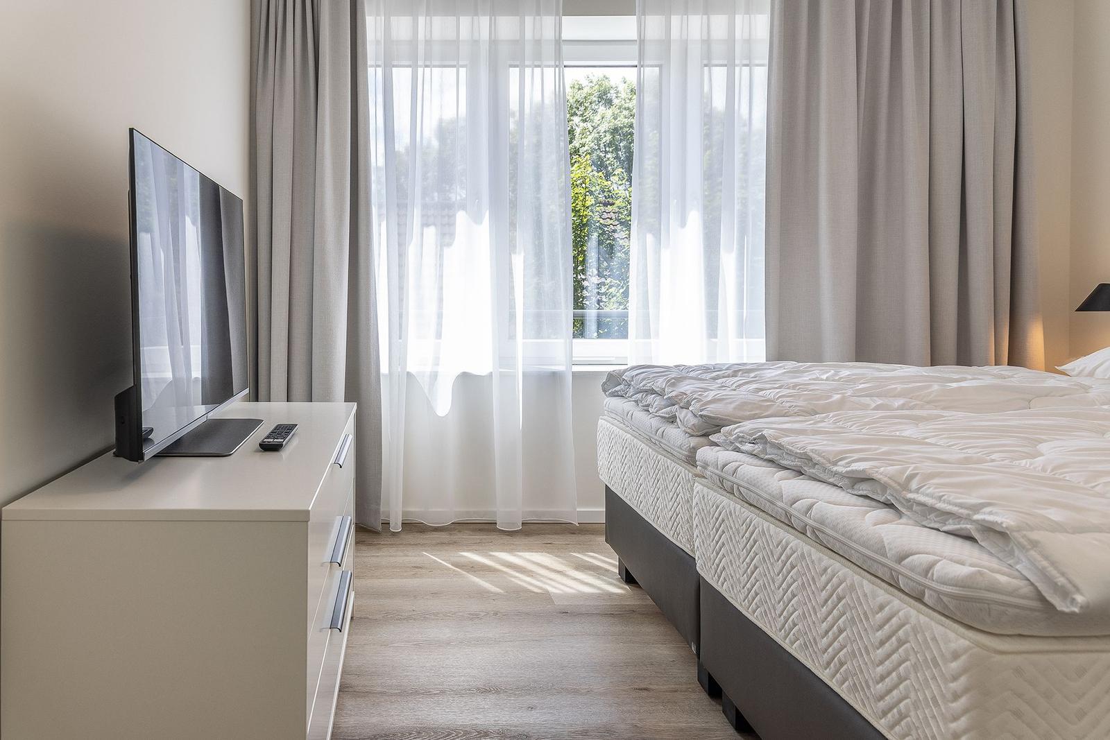 Schlafzimmer mit Doppelbett, TV und Fenster mit Vorhängen.