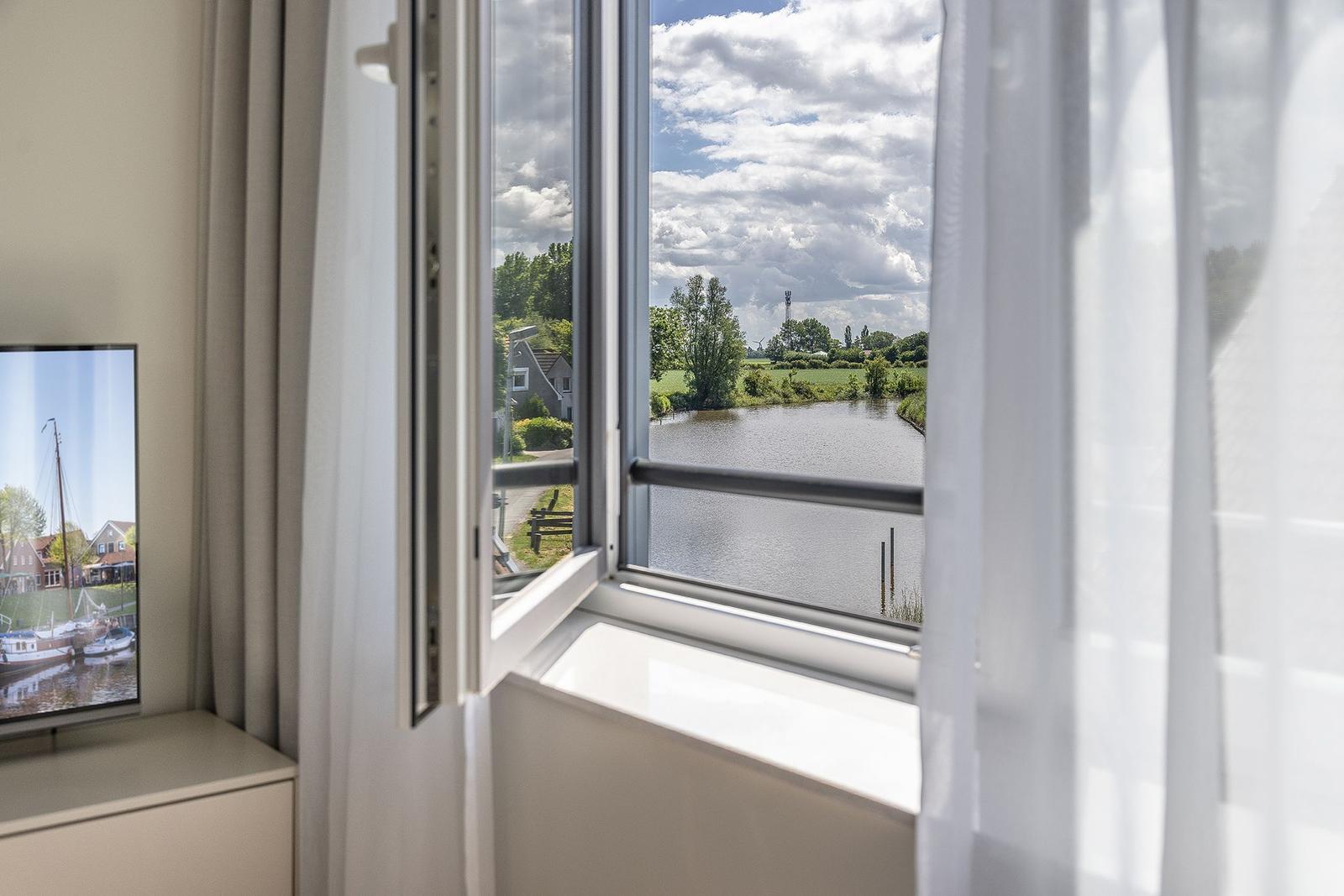 Durch das Fenster blickt man auf einen Kanal mit grünen Ufern und Häusern im Hintergrund.