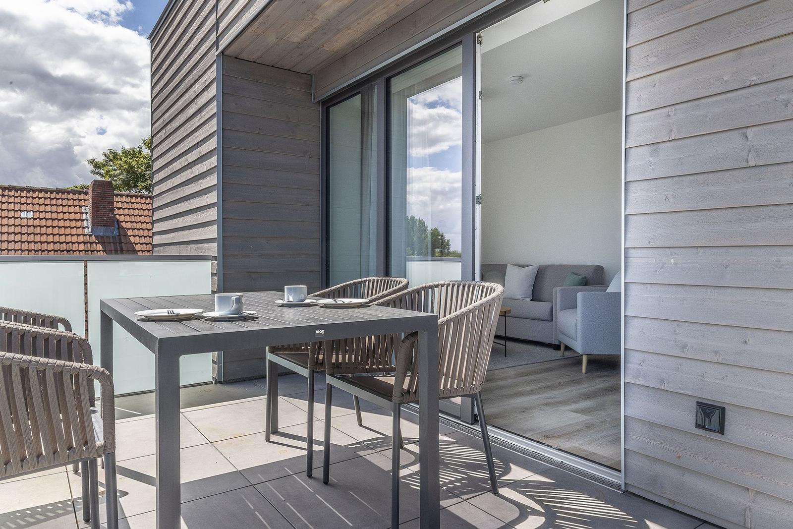 Terrasse mit Tisch und Stühlen. Durch Glastür ist Wohnzimmer mit Sofa sichtbar.