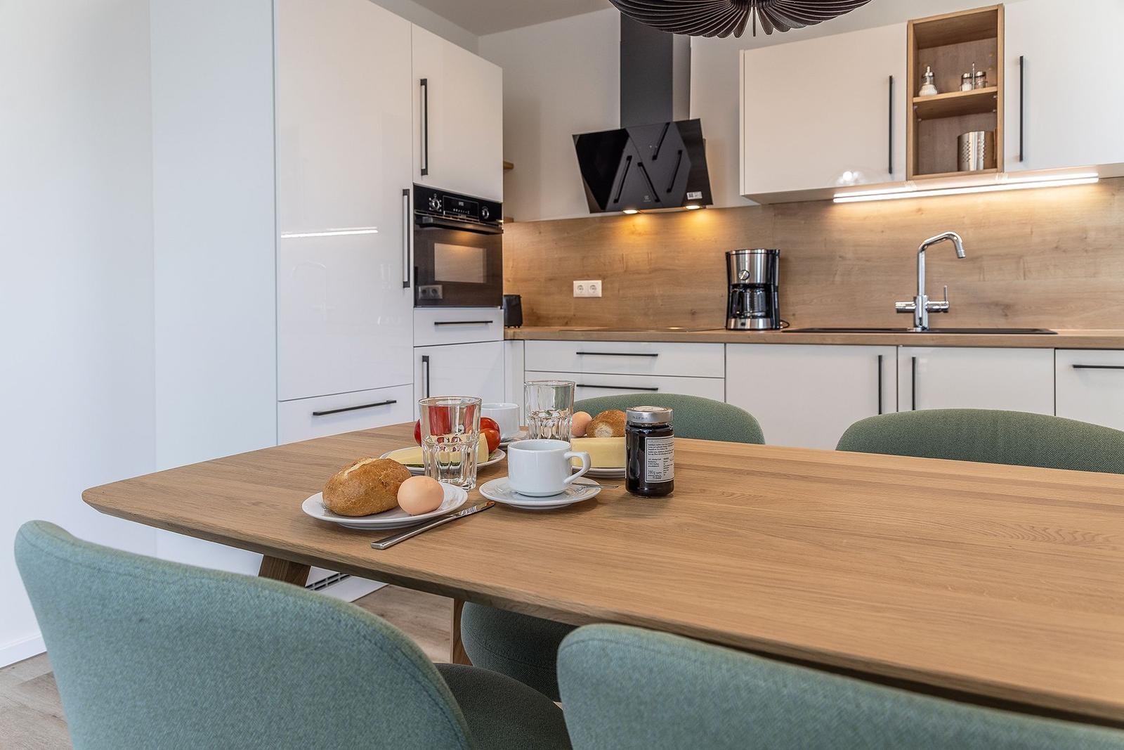 Essbereich mit Holztisch und grünen Stühlen. Küche mit weißen Schränken und Holzrückwand.