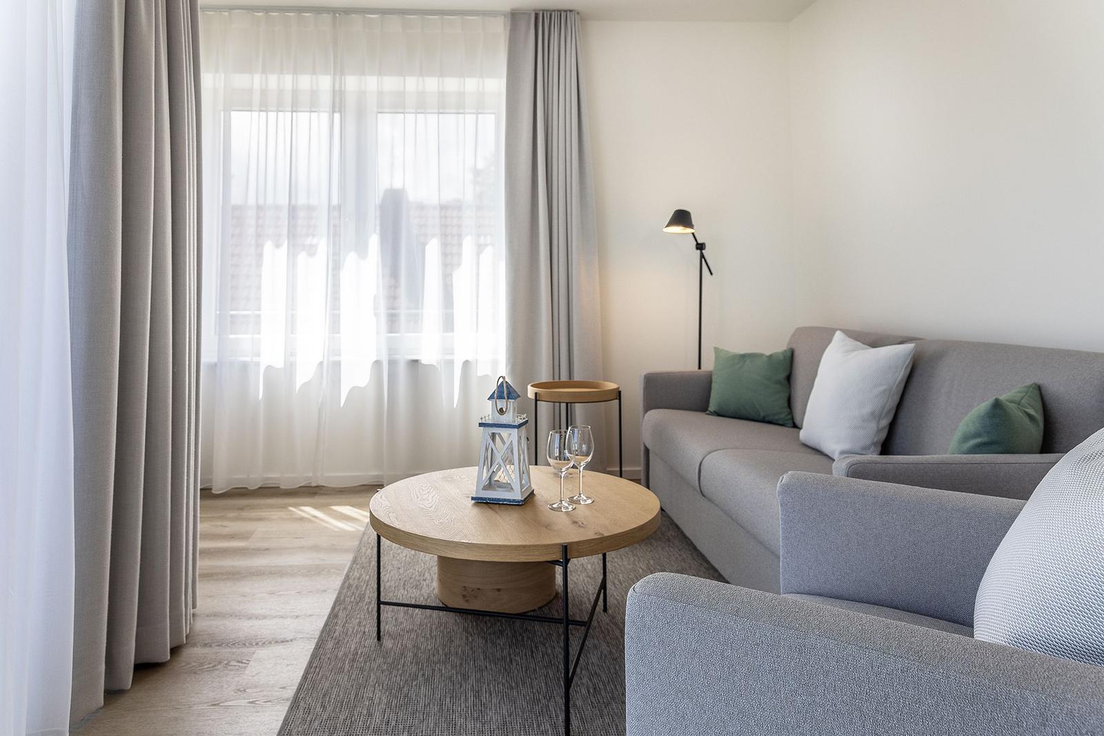 Ein Wohnzimmer mit grauem Sofa, runden Tisch und Fenster mit Vorhängen.