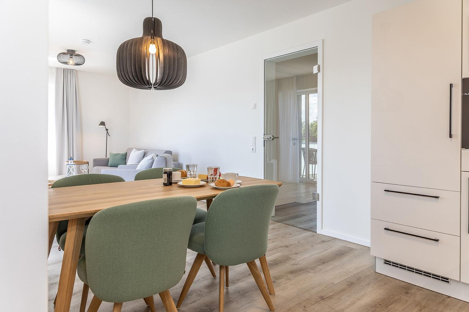 Essbereich mit Holztisch, grünen Stühlen und Küchenzeile. Im Hintergrund Wohnbereich mit Sofa.
