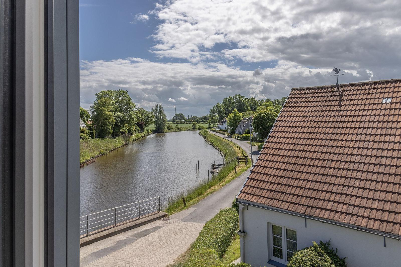 Aus dem Fenster blickt man auf einen Kanal mit Uferweg und Häusern unter bewölktem Himmel.