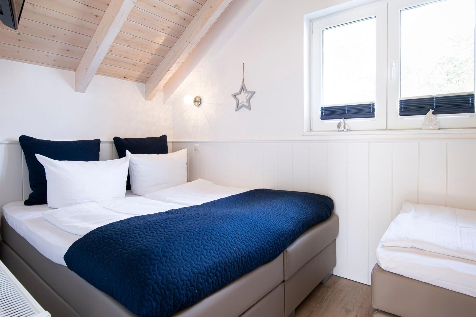 Schlafzimmer mit Doppelbett und kleinem Bett, Holzdecke und Fenster mit Jalousien.