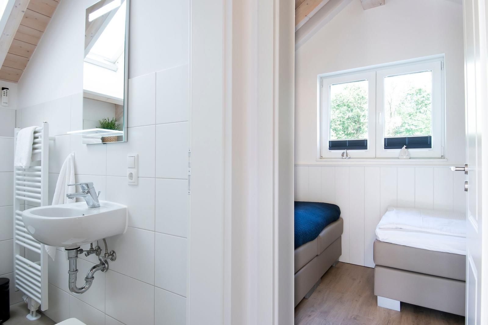 Badezimmer mit Waschbecken und Spiegel. Nebenraum mit zwei Betten und Fenster.