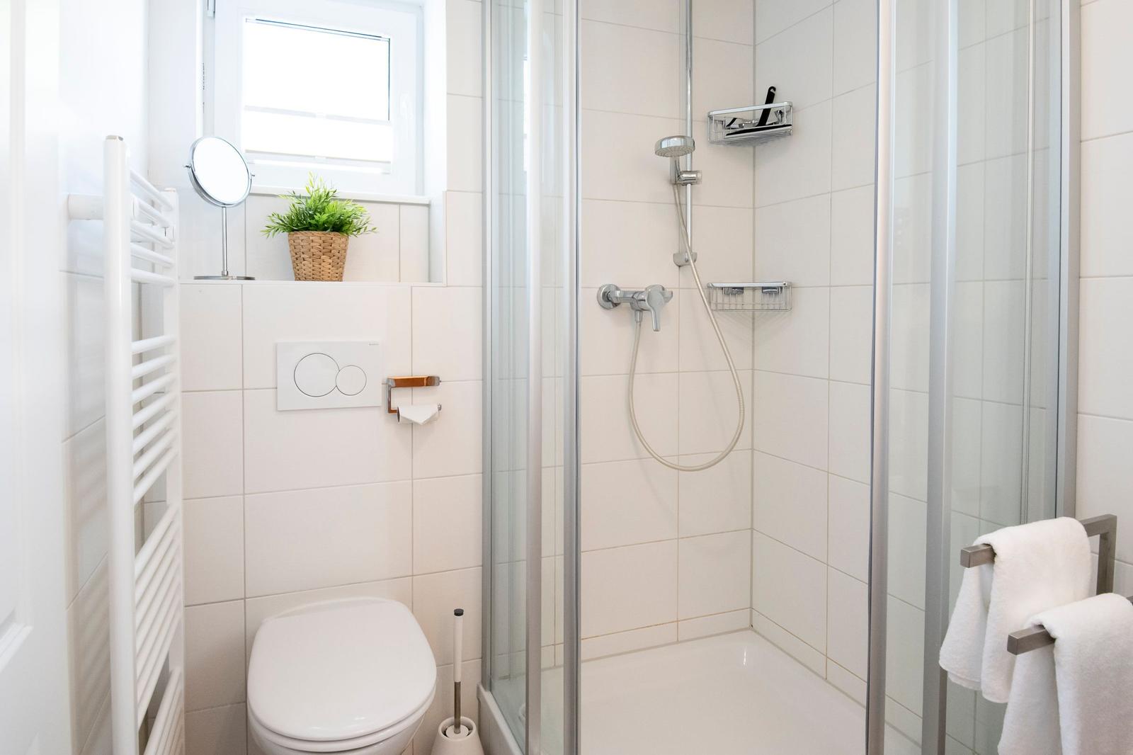 Badezimmer mit Dusche, Toilette und Fenster. Weiße Fliesen, Handtuchhalter und Pflanze.