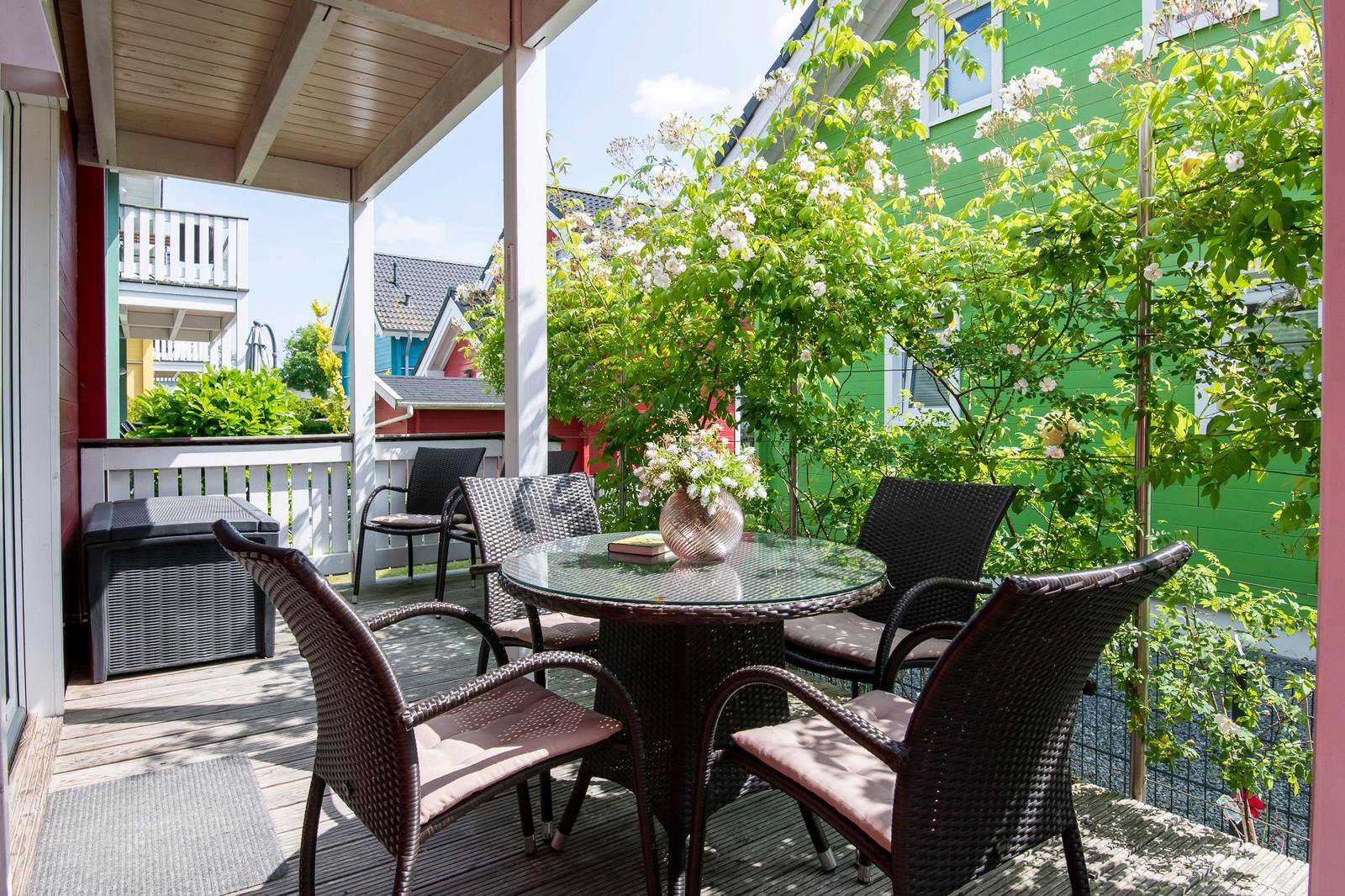 Terrasse mit Tisch und Stühlen, umgeben von grünen Pflanzen und einem grünen Haus.
