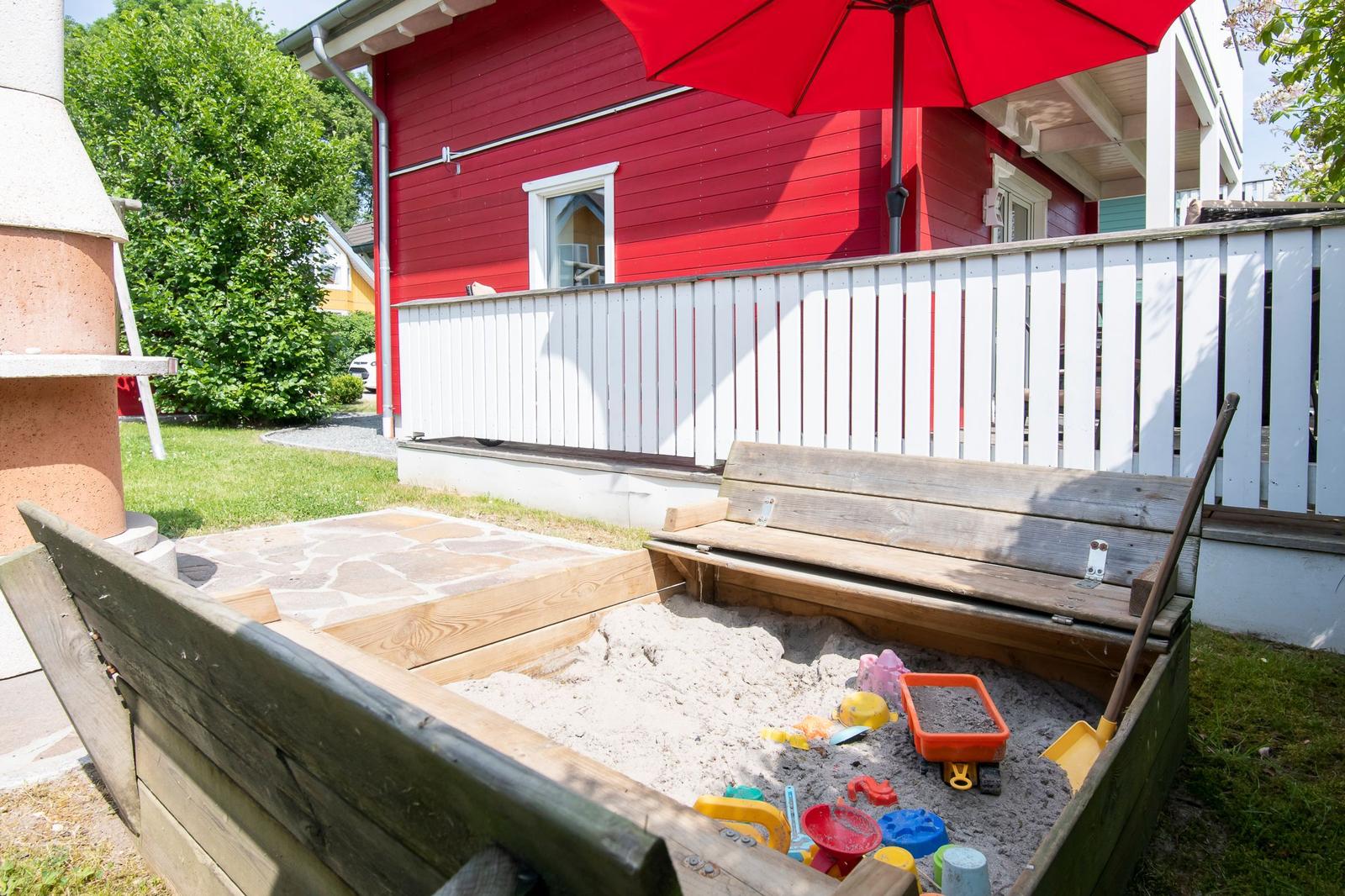 Holzspielplatz mit Sand und bunten Spielzeugen vor rotem Haus mit weißem Zaun.