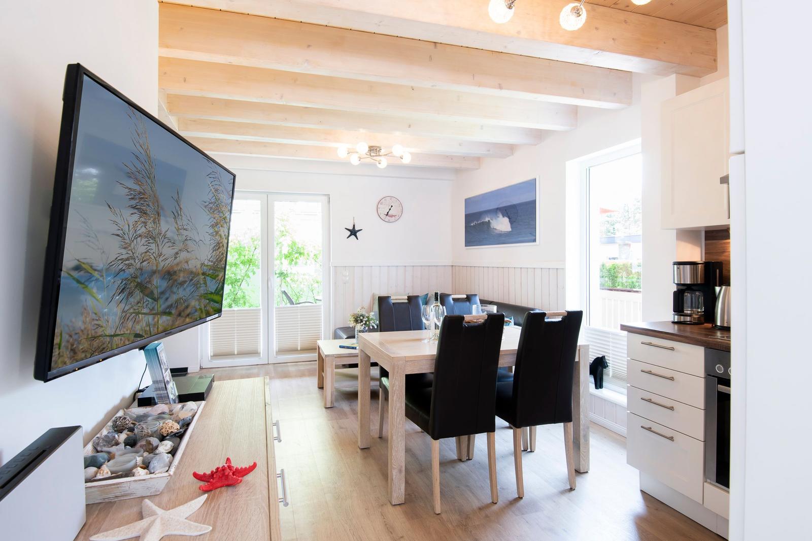 Wohn- und Essbereich mit TV, Esstisch und Küchenzeile. Holzdeckenbalken und Fenster mit Blick.