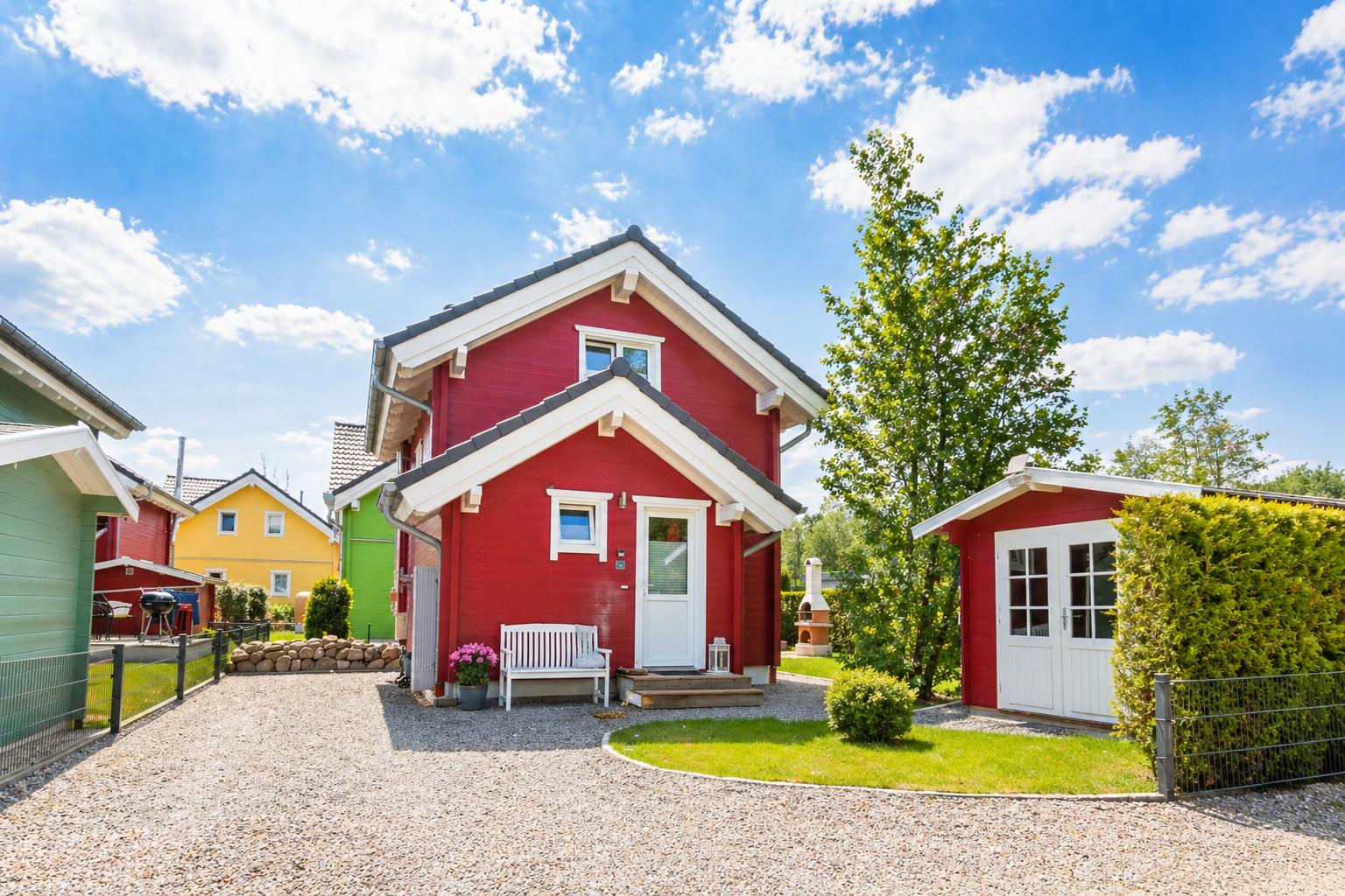Rotes Ferienhaus mit weißer Tür und Bank vorne. Grasfläche und Kiesweg. Nebenhaus mit Gitterzaun.