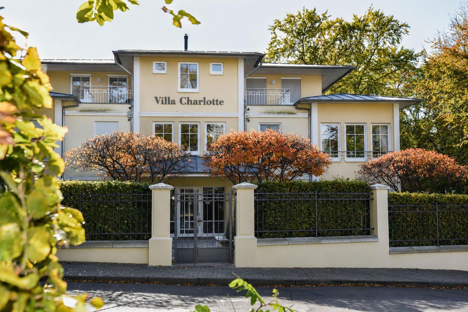Villa Charlotte mit Eingangstor, Hecke und Bäumen im Herbst.