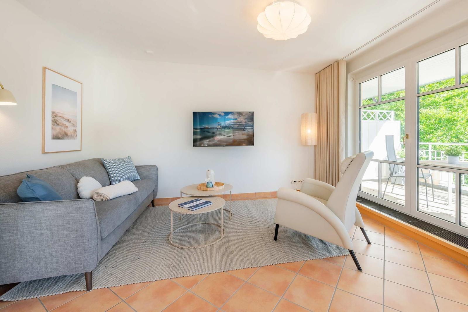 Wohnzimmer mit Sofa, Sessel, Couchtisch und Fernseher. Durchgang zur Terrasse mit Blick auf Garten.