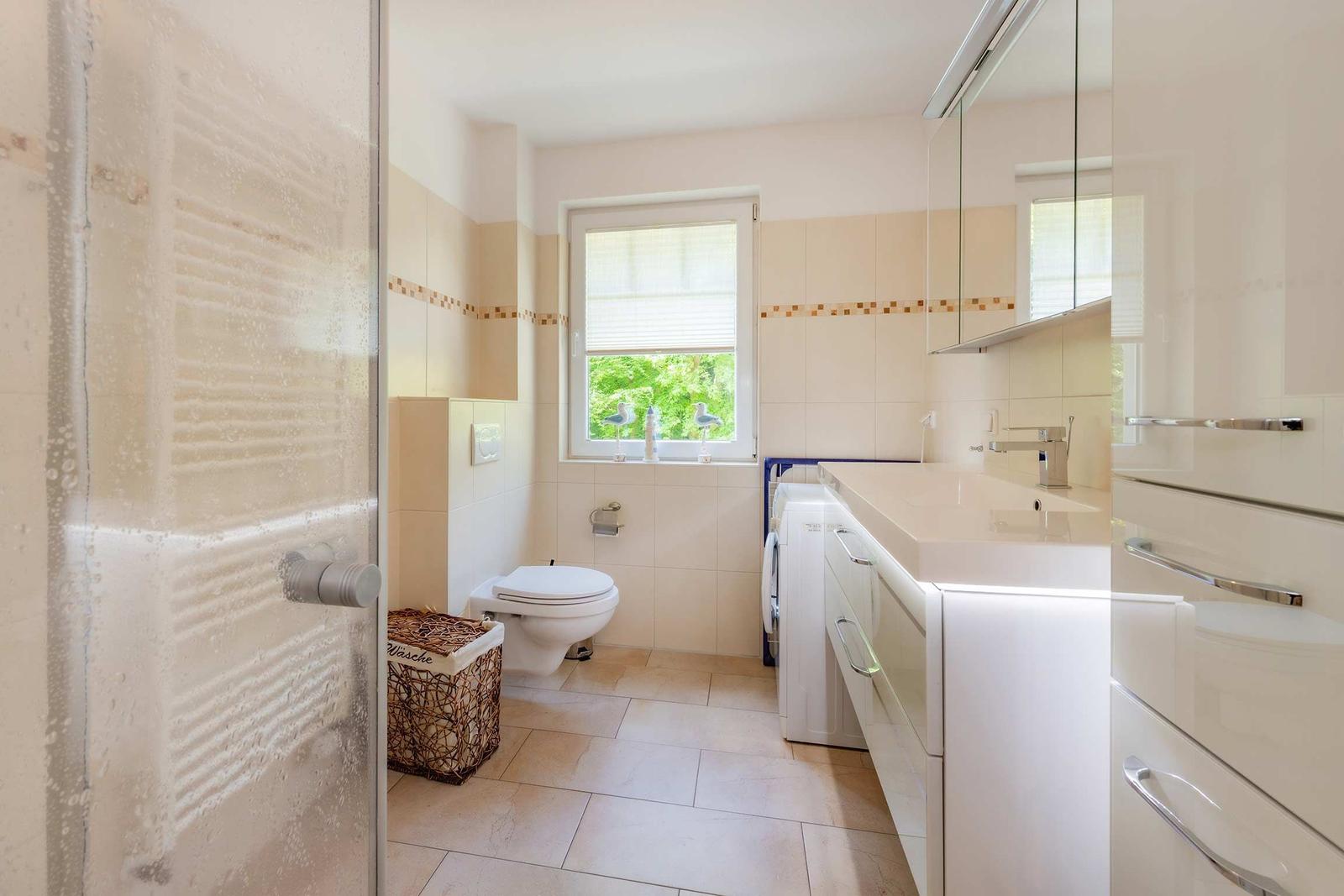 Badezimmer mit Dusche, Toilette, Waschmaschine und Fenster mit Blick auf Grün.