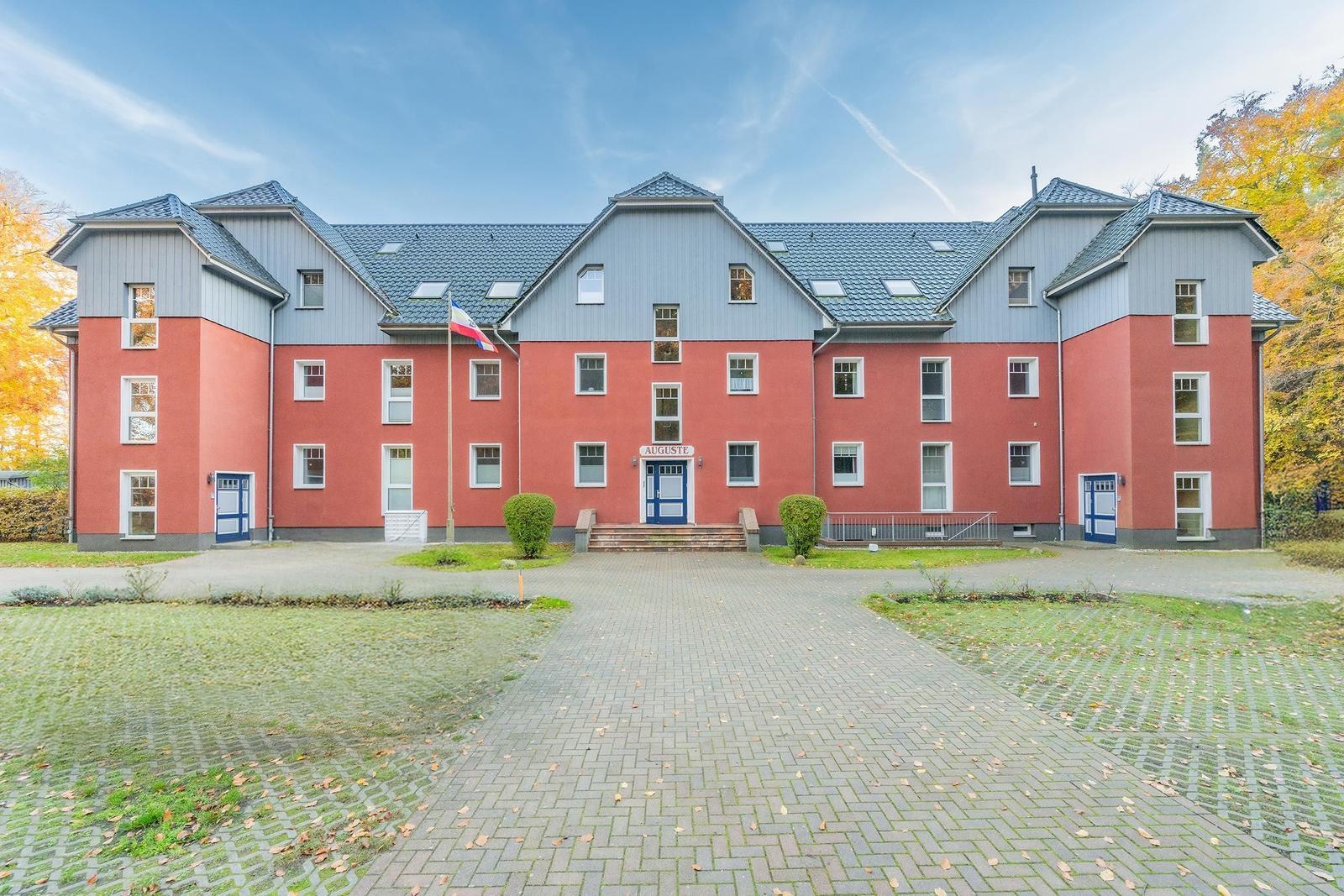 Rotes Gebäude mit blauem Dach und Eingangstür. Vorhof mit gepflastertem Weg und Bäumen.