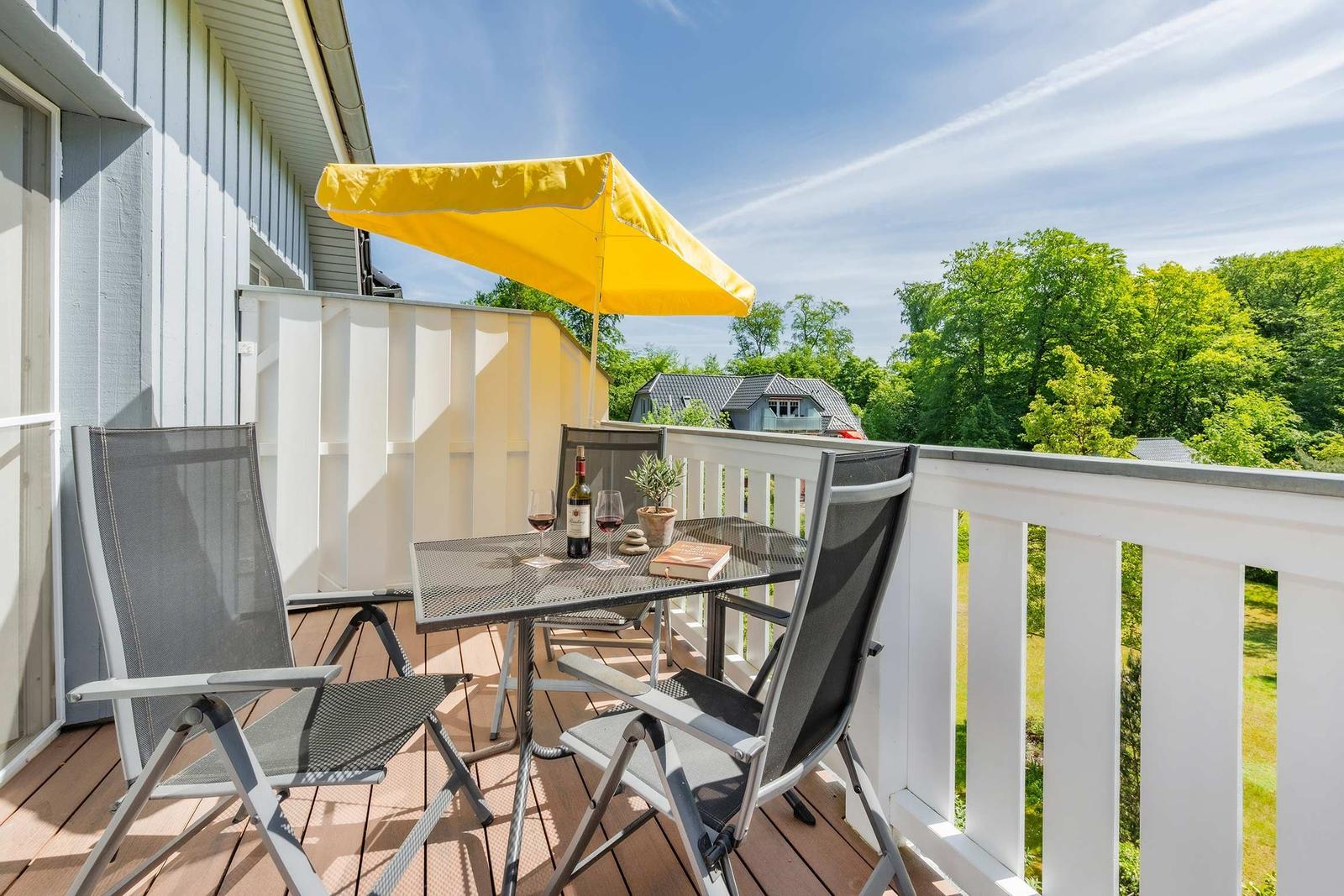 Terrasse mit Tisch, Stühlen und gelbem Sonnenschirm. Blick auf grüne Bäume und einen Nachbarhof.