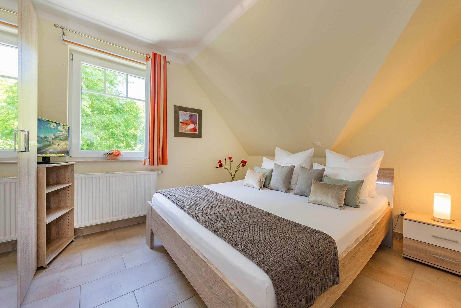 Schlafzimmer mit Dachschrägen, Bett, Nachttisch, TV und Fenster mit Blick auf Grün.