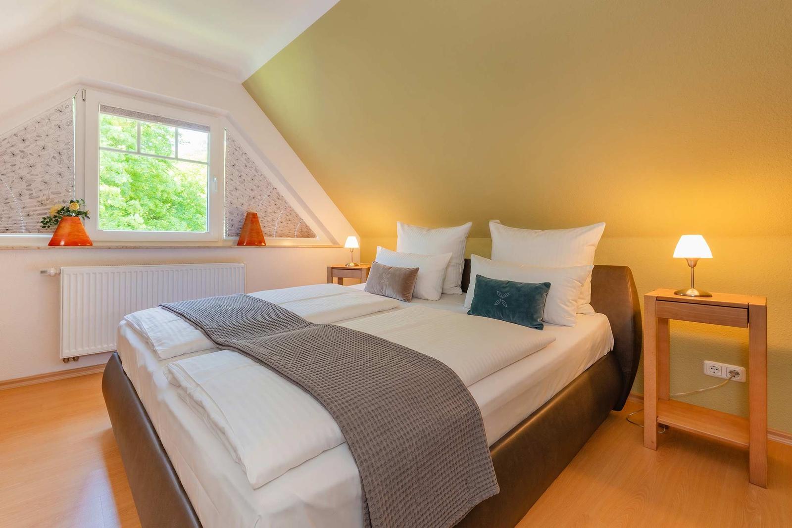 Doppelbett mit Bettwäsche, Nachttisch und Fenster mit Blick auf Bäume.