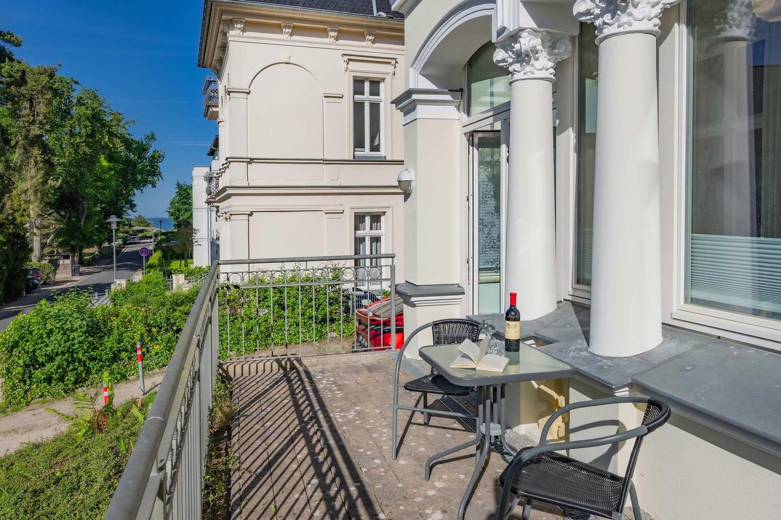Terrasse mit Tisch, Stühlen und Flasche. Blick auf Straße und Grünflächen.