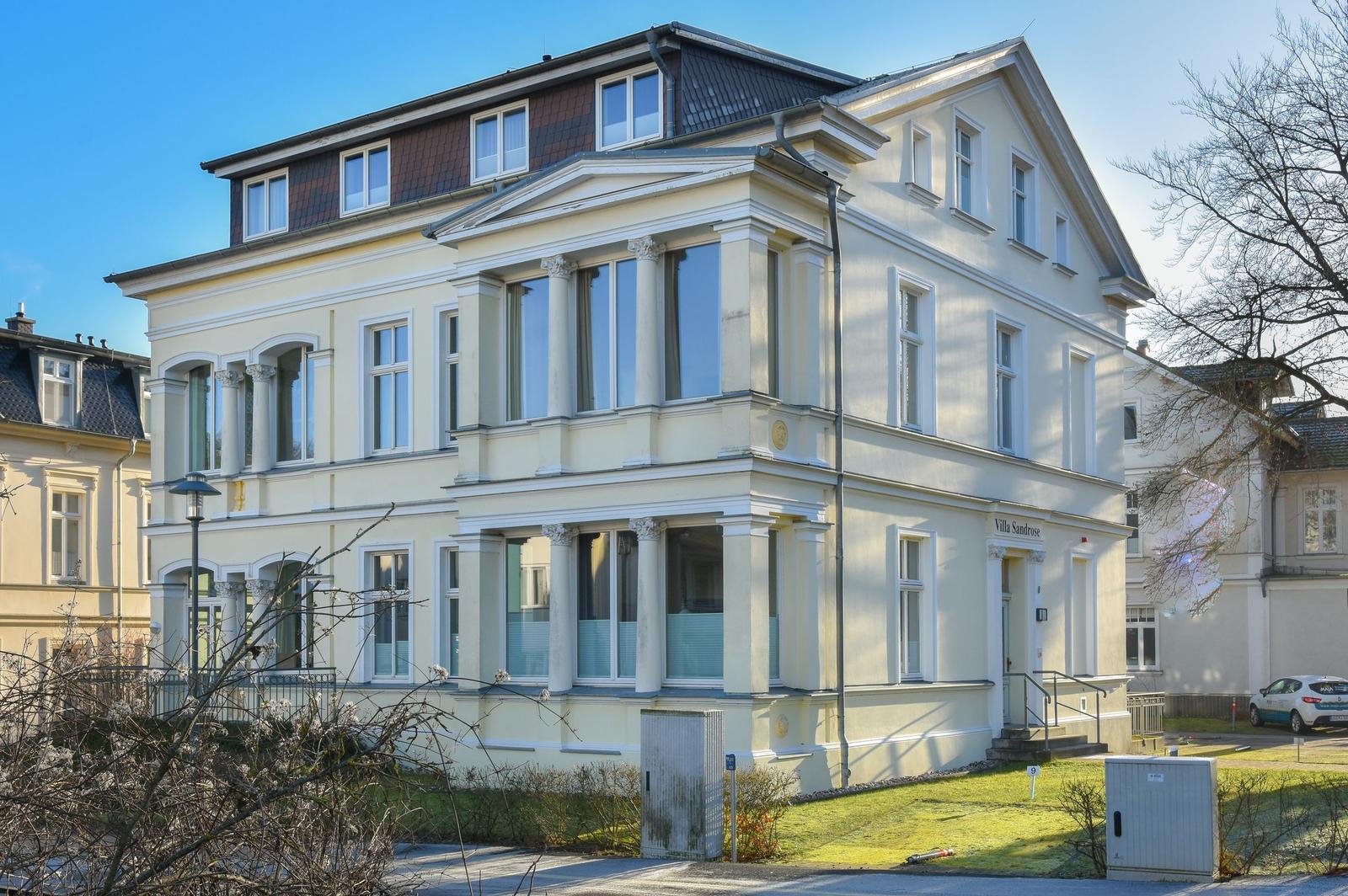 Villa Sandrose mit hellen Fassaden und Säulen. Vorderhof mit Rasen und Gehweg.