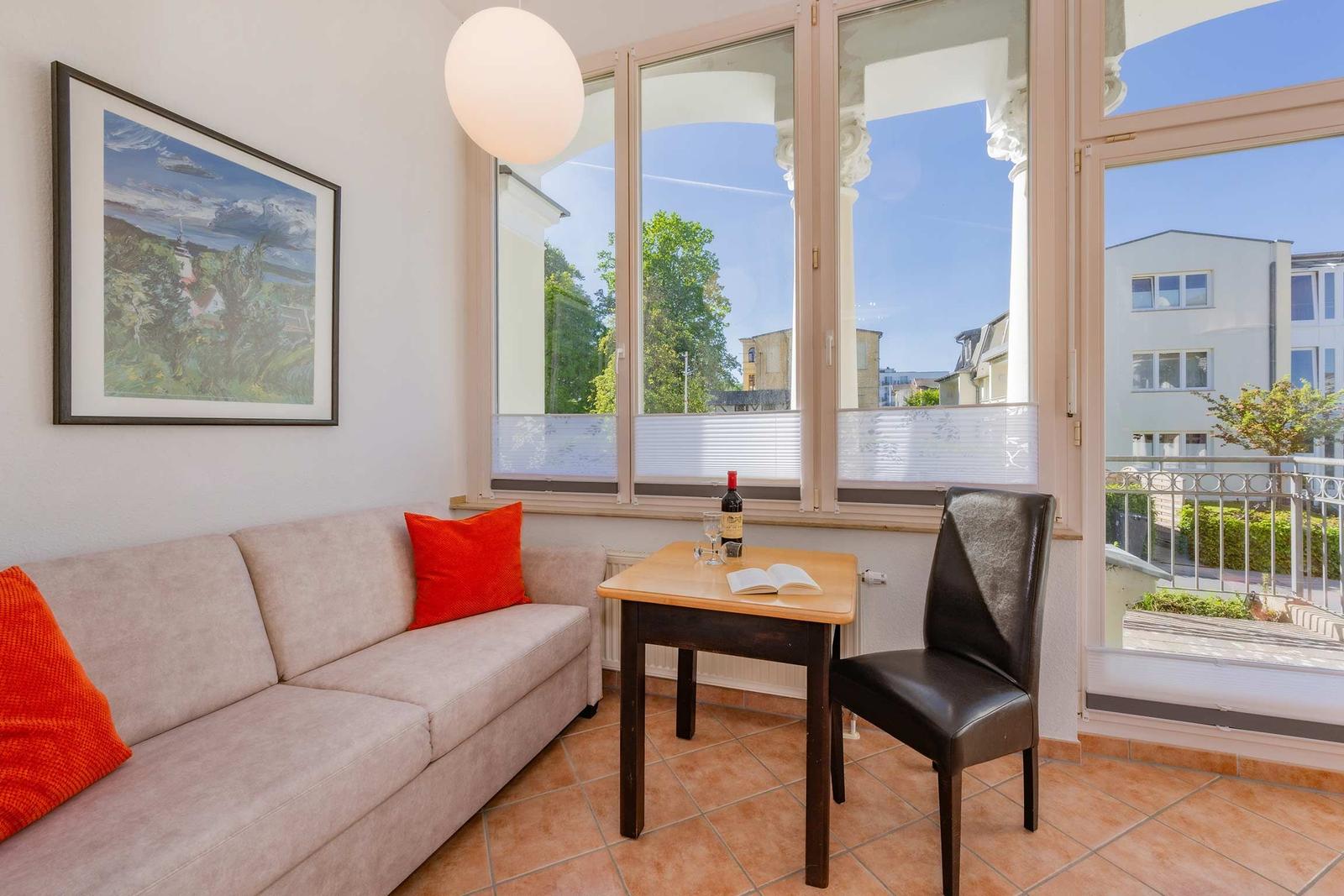 Wohnbereich mit Sofa, Tisch und Stuhl. Große Fenster mit Blick auf Balkon und Umgebung.