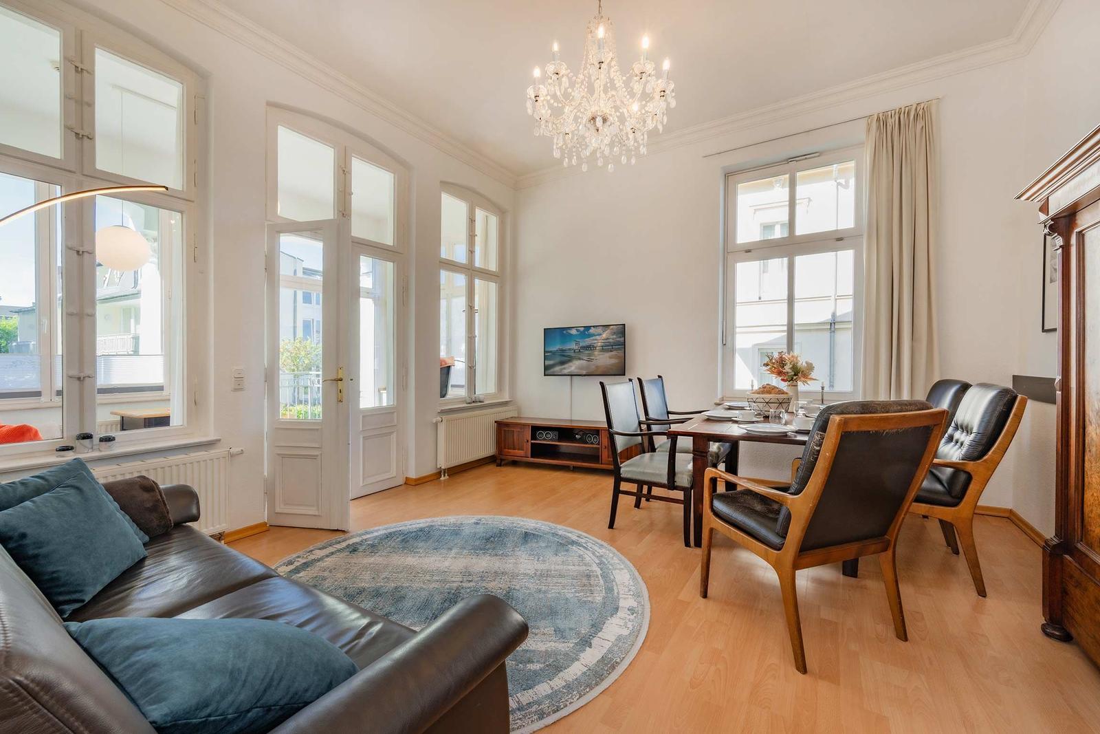Wohn- und Essbereich mit Sofa, Esstisch und Kronleuchter. Holzboden und große Fenster.
