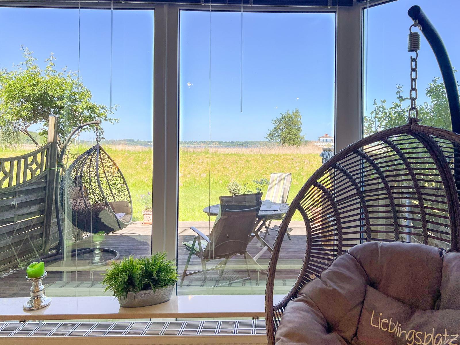 Rattan-Schaukelstühle vor großem Fenster mit Blick auf Garten und Landschaft.